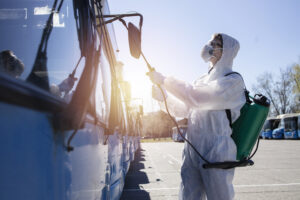 Public transport disinfection. Man in white protective suit with reservoir spraying disinfectant on parked buses. Stop coronavirus or COVID-19.